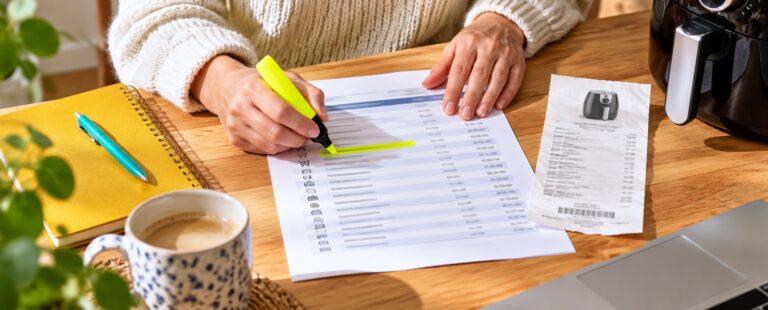 Personne contrôlant des prélèvements récurrents sur un relevé bancaire près d’un ticket d’achat d’électroménager.